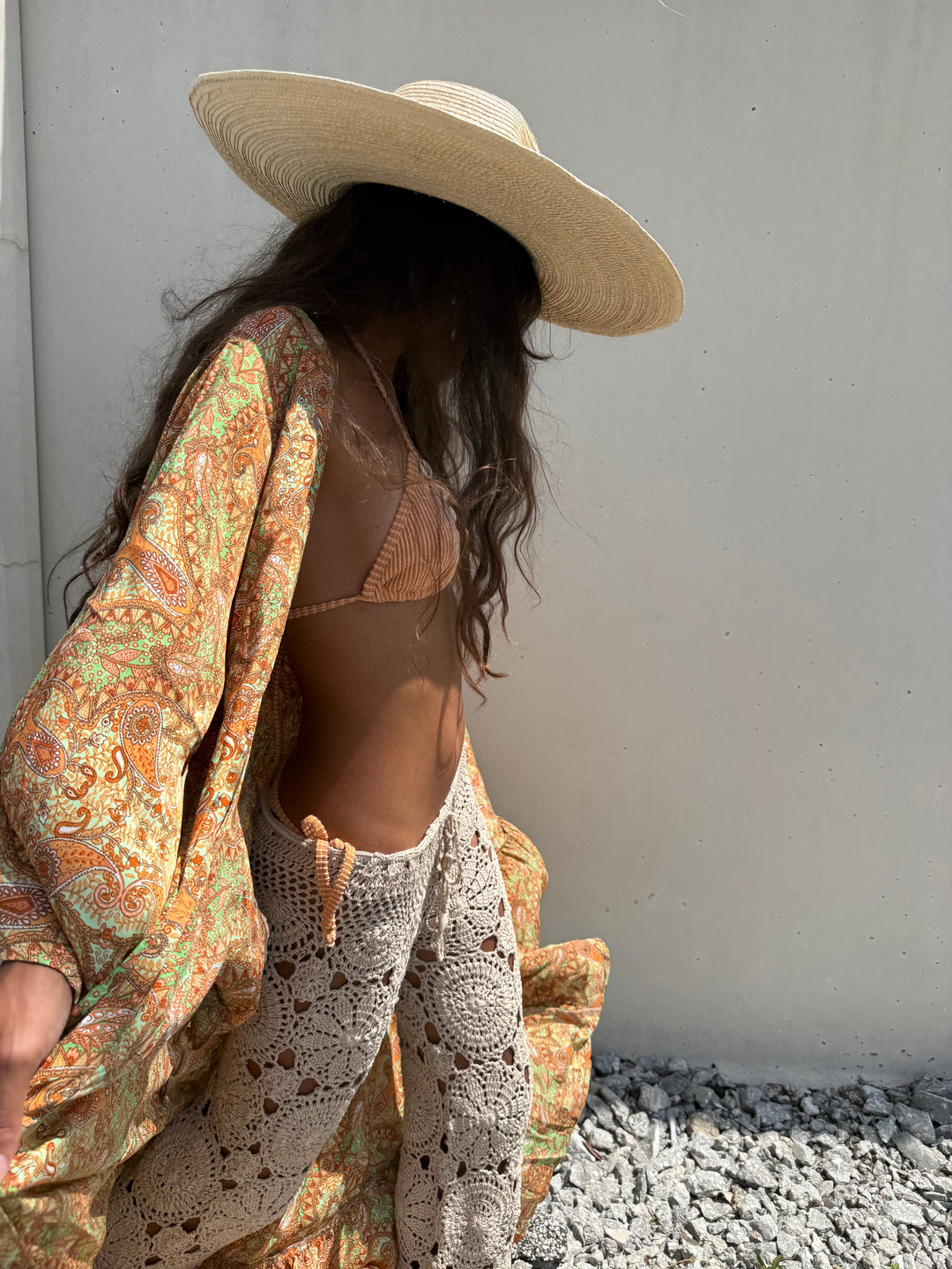 A woman wearing a wide-brimmed straw hat, a tan crochet bikini, a loose paisley orange and green cover-up, and white crochet pants, standing against a plain light gray wall.