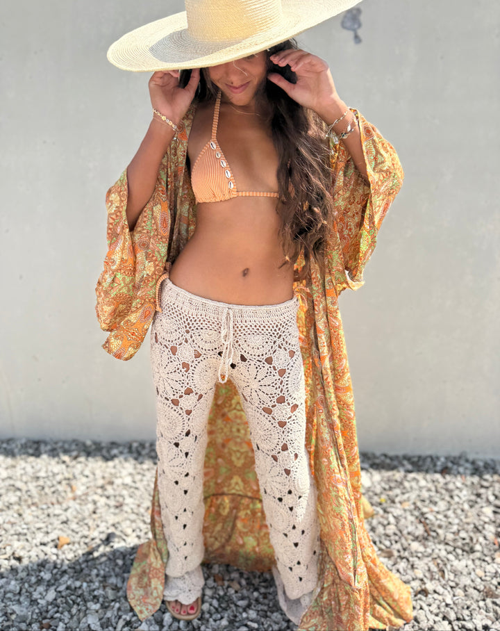 A woman wearing a bohemian-style outfit featuring a wide-brimmed straw hat, a peach bikini top, a yellow and orange patterned open-front kaftan, and cream crochet pants, standing outdoors against a light gray wall.
