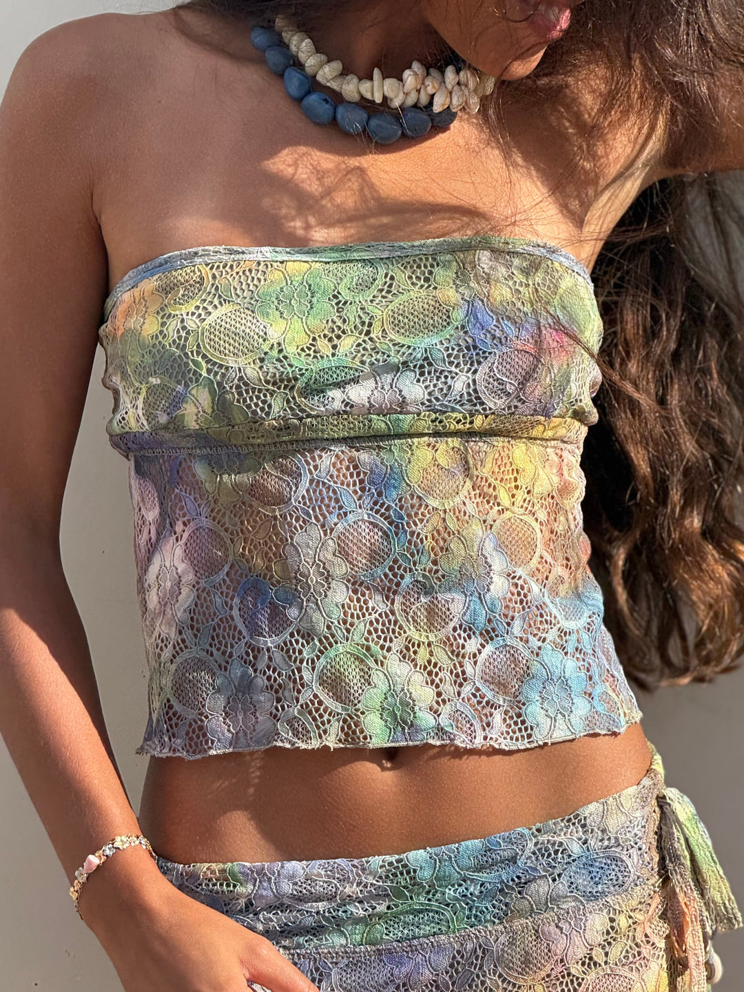 A woman wearing a matching lace two-piece set with a strapless bandeau top and coordinating skirt or sarong, featuring a multicolor pastel pattern in green, blue, yellow, and lavender. She is also wearing a blue and white beaded necklace and a silver bracelet.