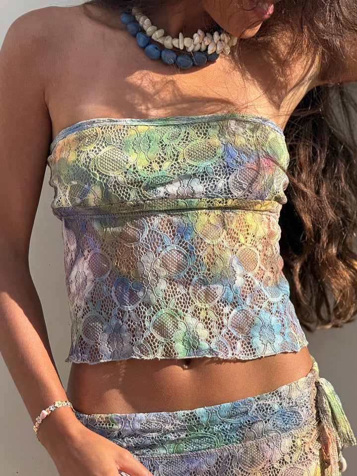 A woman wearing a matching lace two-piece set with a strapless bandeau top and coordinating skirt or sarong, featuring a multicolor pastel pattern in green, blue, yellow, and lavender. She is also wearing a blue and white beaded necklace and a silver bracelet.