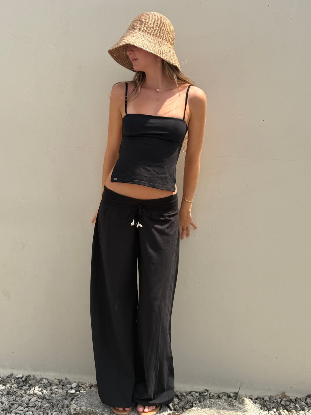 A woman wearing a straw bucket hat, a black spaghetti-strap crop top, and black wide-leg pants with white drawstrings, standing against a plain beige wall.