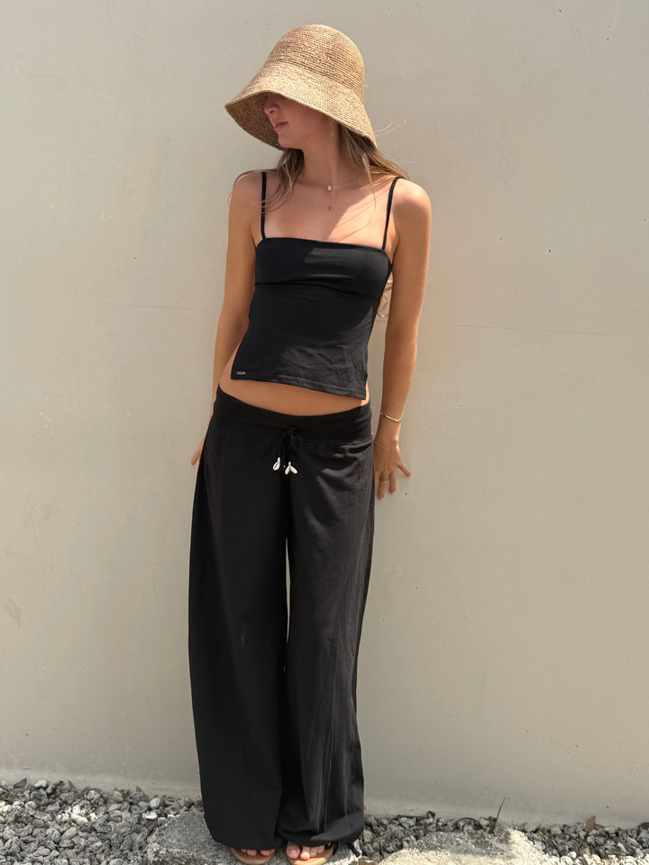 A woman wearing a straw bucket hat, a black spaghetti-strap crop top, and black wide-leg pants with white drawstrings, standing against a plain beige wall.