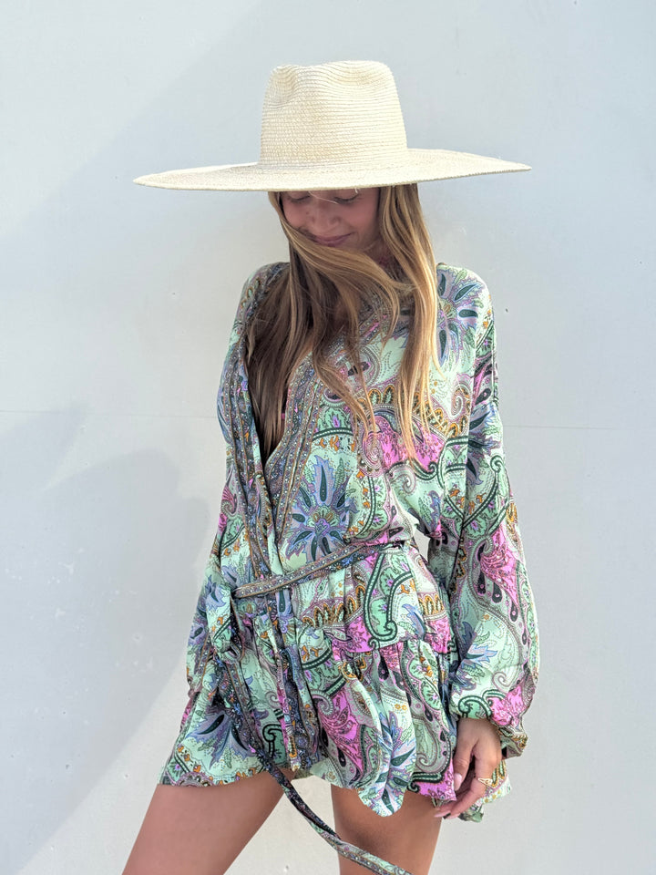 A woman wearing a wide-brimmed straw hat and a long-sleeve paisley romper with a multicolor floral and paisley print in green, pink, blue, and beige, standing against a plain light gray wall.