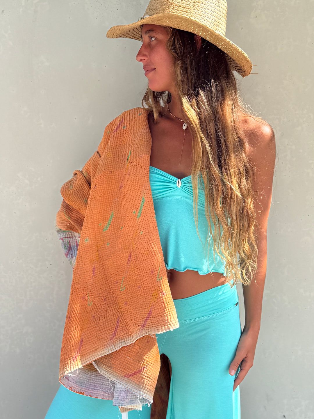 A woman wearing a straw hat, a turquoise two-piece set, and an orange textured wrap draped over her shoulder, standing against a light gray wall.