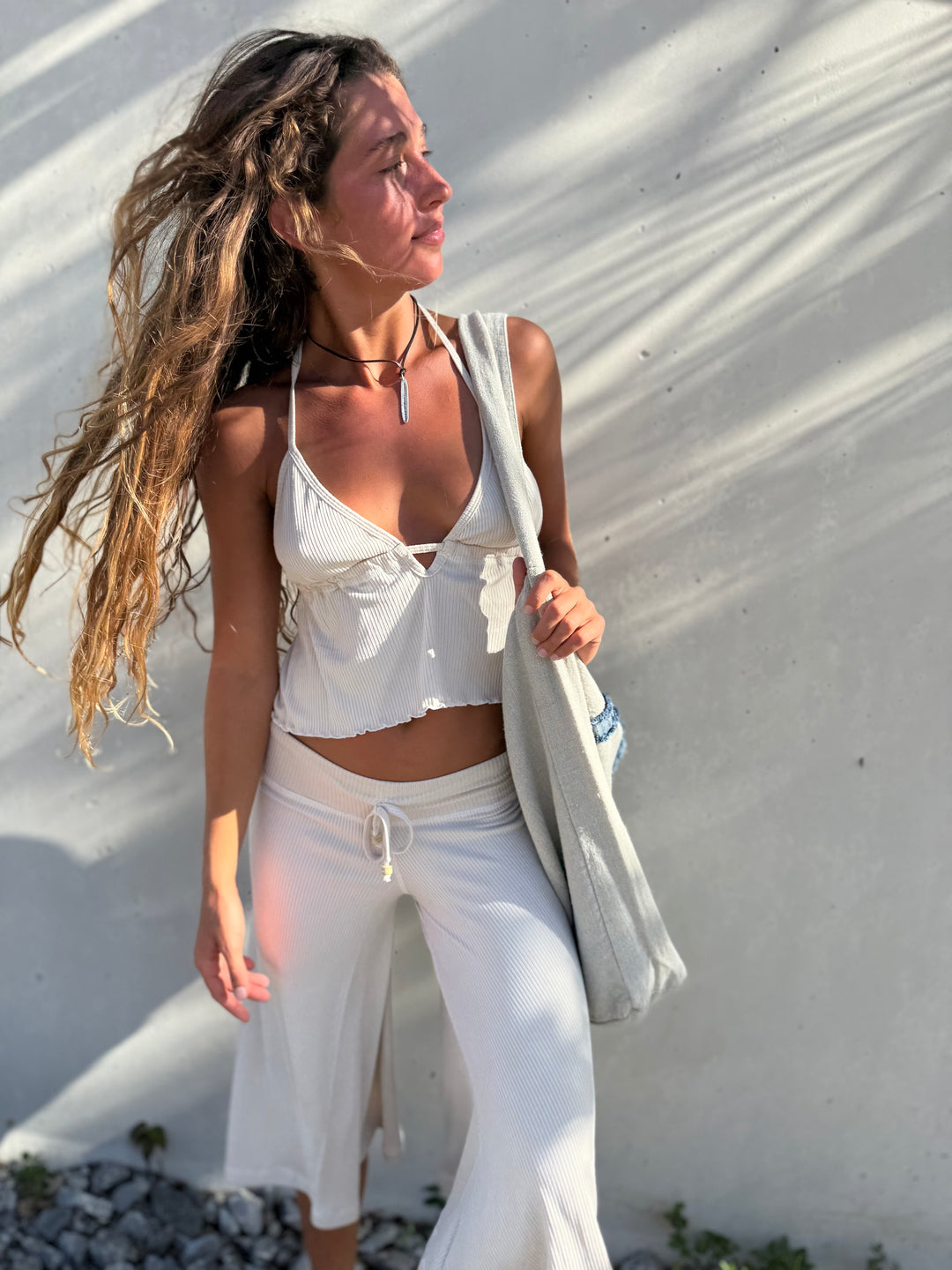 A woman stands outdoors against a sunlit white wall wearing a white cropped camisole, white wide-leg pants, and carrying a light gray tote bag.