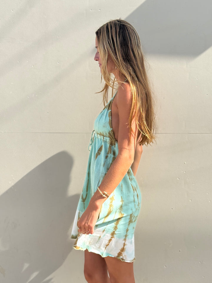 A woman wearing a light blue and white tie-dye halter dress stands in profile against a sunlit beige wall.