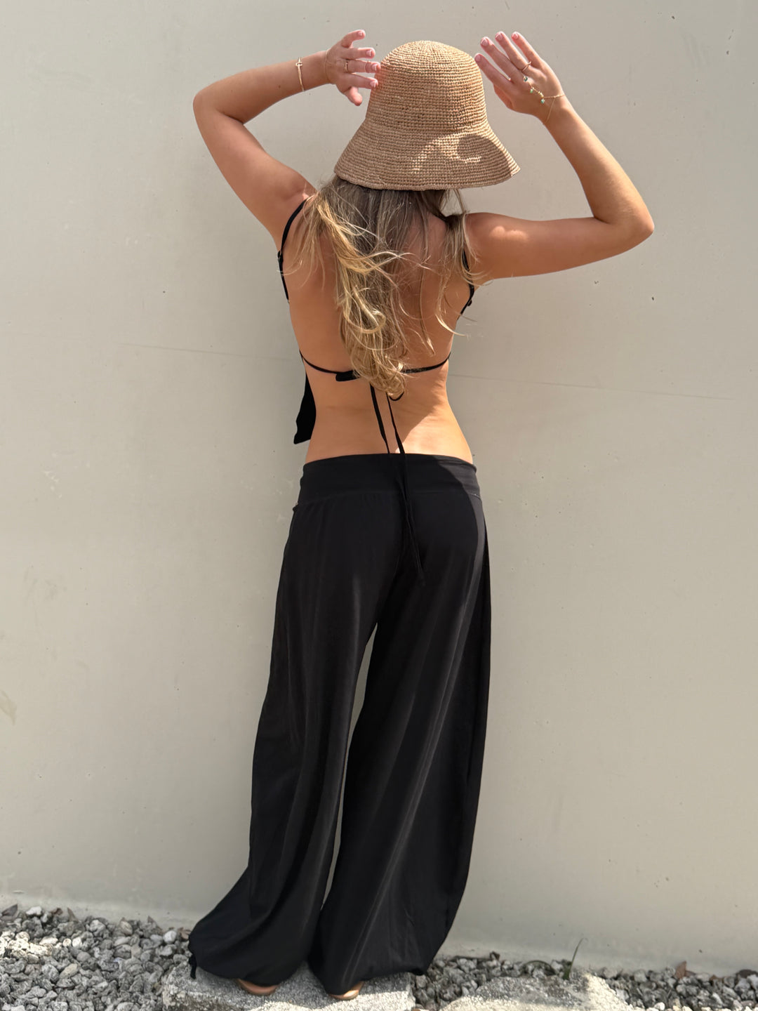 A woman stands with her back to the camera wearing a beige straw bucket hat, a black strappy bikini top, and loose black wide-leg pants against a plain light wall.