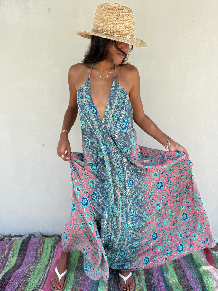 A woman wearing a straw hat and sunglasses models a sleeveless halter maxi dress with a blue and pink paisley pattern, holding out the sides of the dress to show its flow. She is standing on a colorful striped rug against a textured off-white wall.