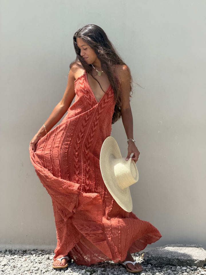 A woman wearing a rust-orange textured maxi dress and holding a beige wide-brim straw hat, standing against a plain light gray wall on a gravel surface.
