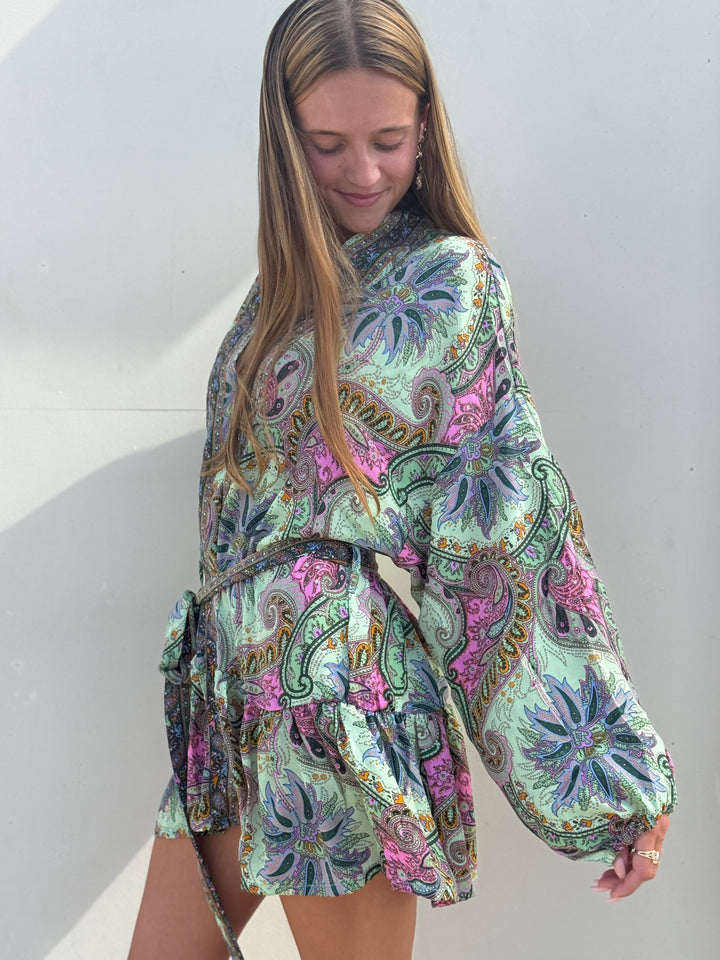 A woman wearing a long-sleeve mini dress with a colorful paisley pattern in shades of green, pink, purple, and blue, standing against a plain light gray wall.