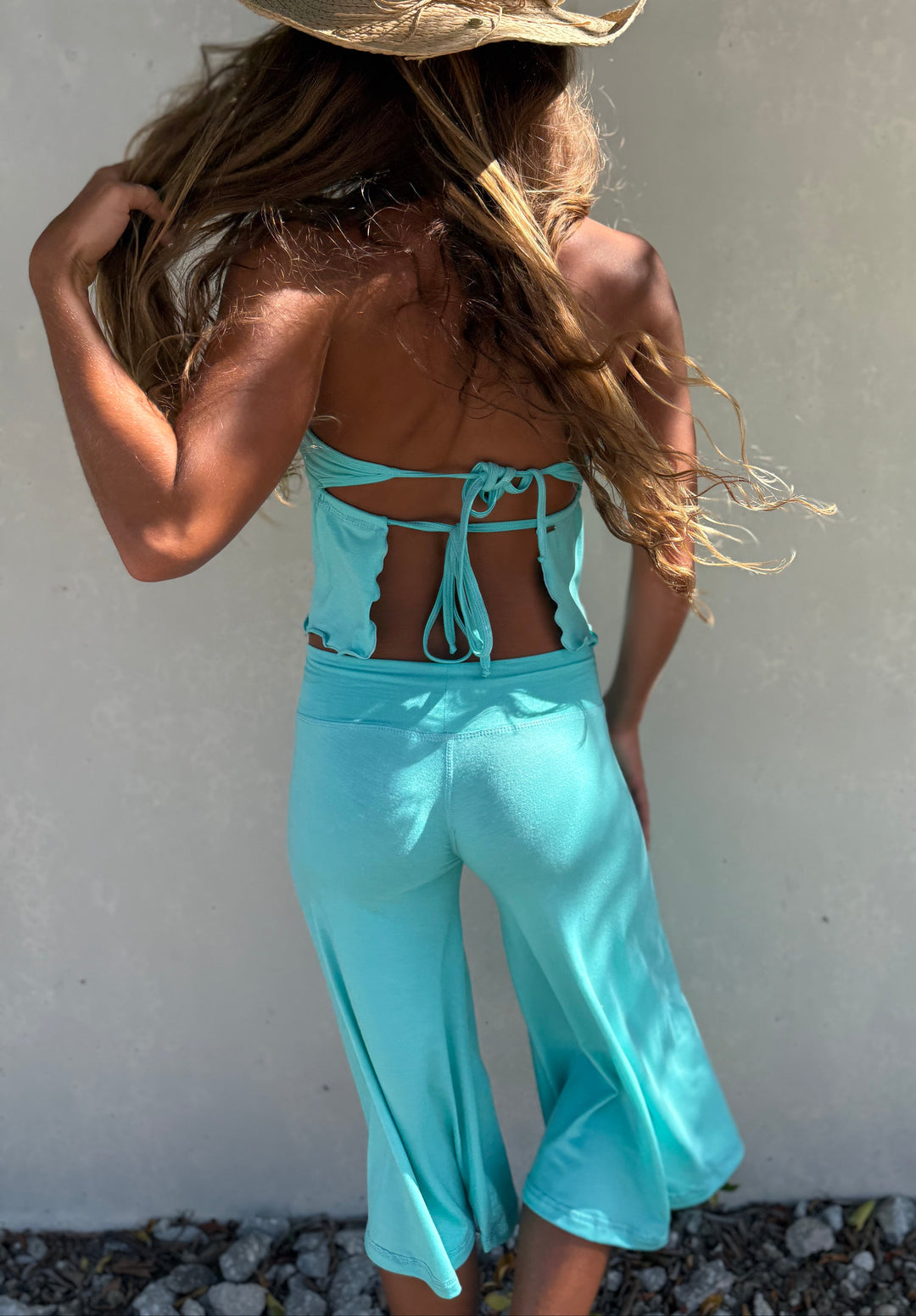A woman wearing a light blue two-piece set with a halter top and matching wide-leg pants, paired with a straw hat, photographed from behind against a light gray wall.