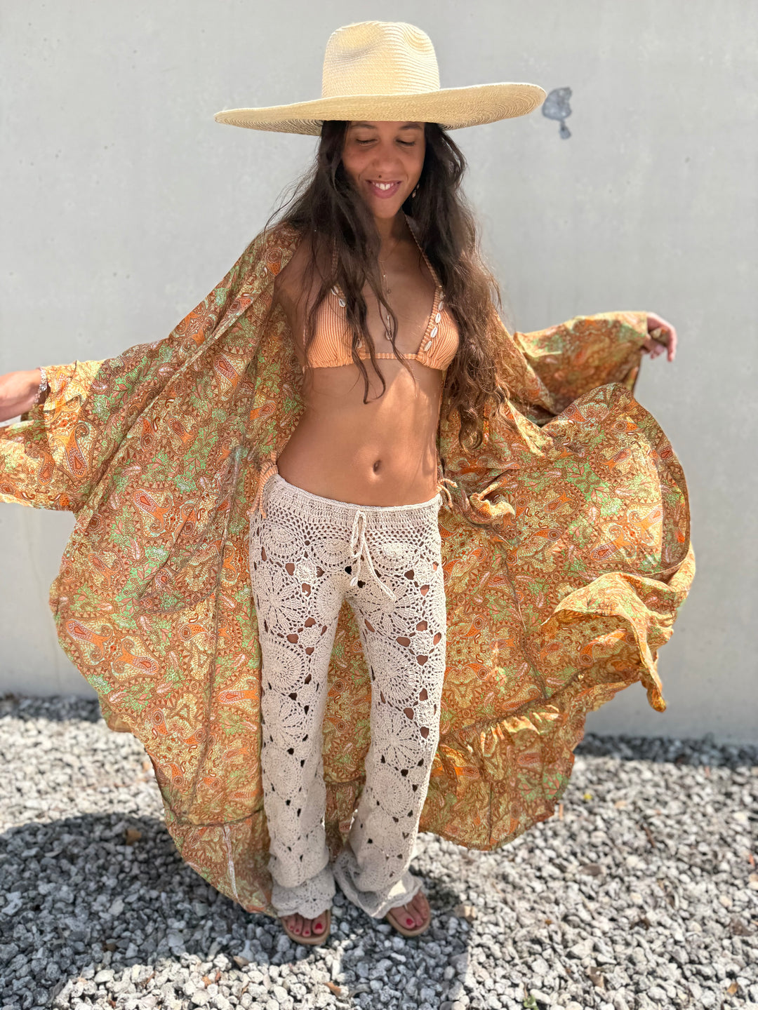 A woman wearing a wide-brim straw hat, a beige bikini, cream crochet pants, and a flowing orange and green paisley-print cover-up stands outdoors against a light gray wall on a gravel surface.