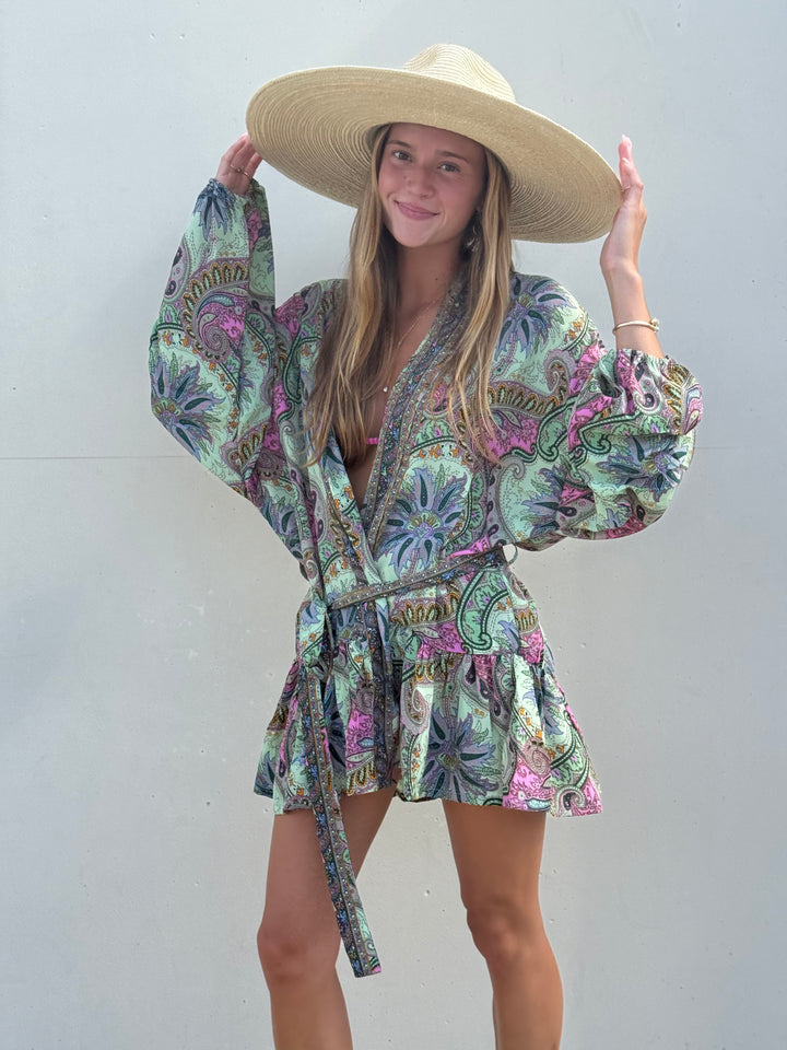 A woman wearing a light beige wide-brim straw hat and a green paisley print wrap robe with pink and purple accents, posing against a plain light gray wall.
