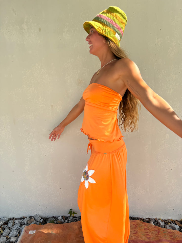 A woman wearing a bright orange strapless dress with a white flower applique and a yellow striped bucket hat, standing against a textured beige wall.