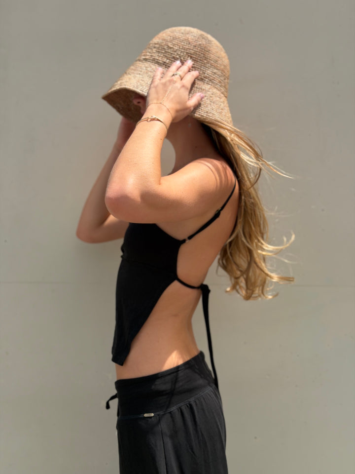 A woman wearing a black strappy cutout camisole top and black bottoms, holding a beige straw bucket hat against her face, set against a plain gray wall.