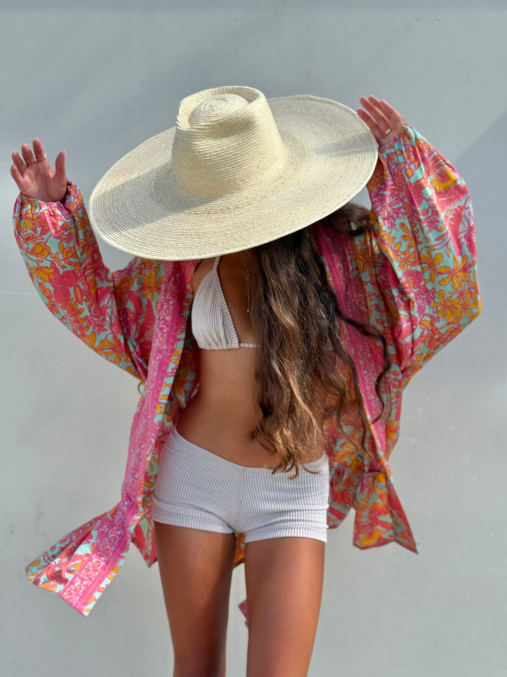 A woman wearing a large straw sun hat, a vibrant pink and multicolor patterned long-sleeve cover-up, and white ribbed bikini shorts and a white bikini top, posing against a light gray wall.