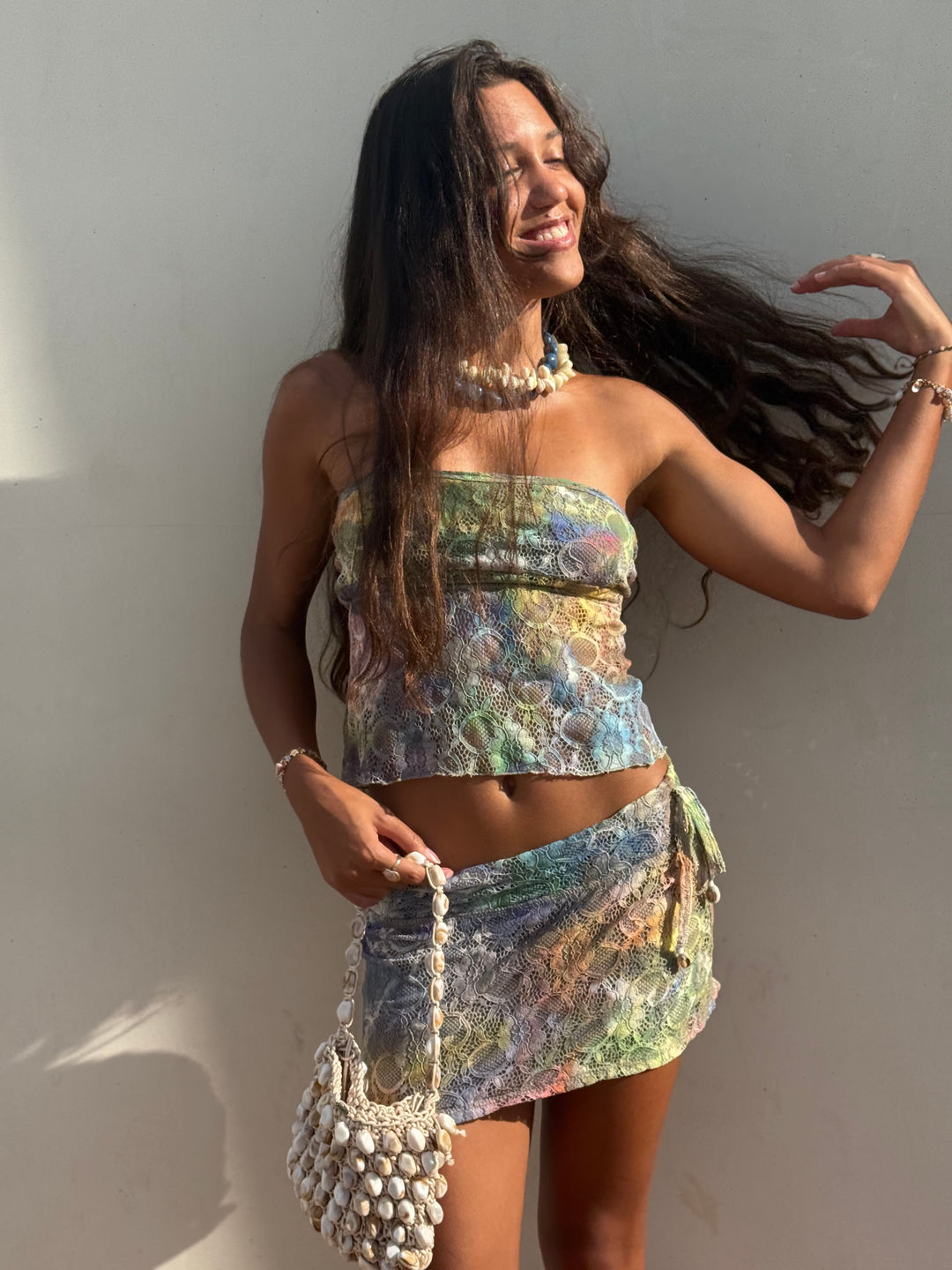 A woman posing in a matching two-piece set featuring a strapless tube top and a wrap skirt, both in a multicolor abstract pattern. She is accessorized with a chunky pearl necklace and a matching beaded handbag, set against a plain light gray wall.