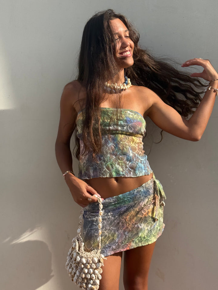 A woman posing in a matching two-piece set featuring a strapless tube top and a wrap skirt, both in a multicolor abstract pattern. She is accessorized with a chunky pearl necklace and a matching beaded handbag, set against a plain light gray wall.