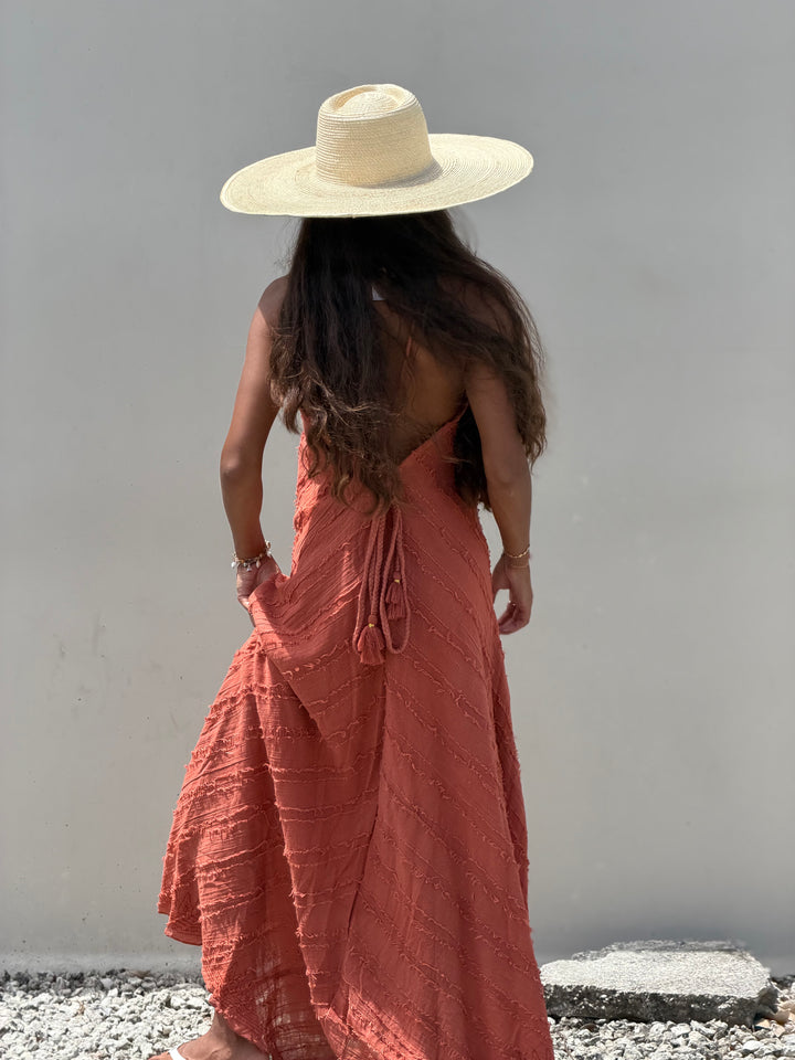 A woman viewed from behind wearing a wide-brimmed straw hat and a rust-orange textured maxi dress with a tie-back detail, standing against a plain light gray wall.