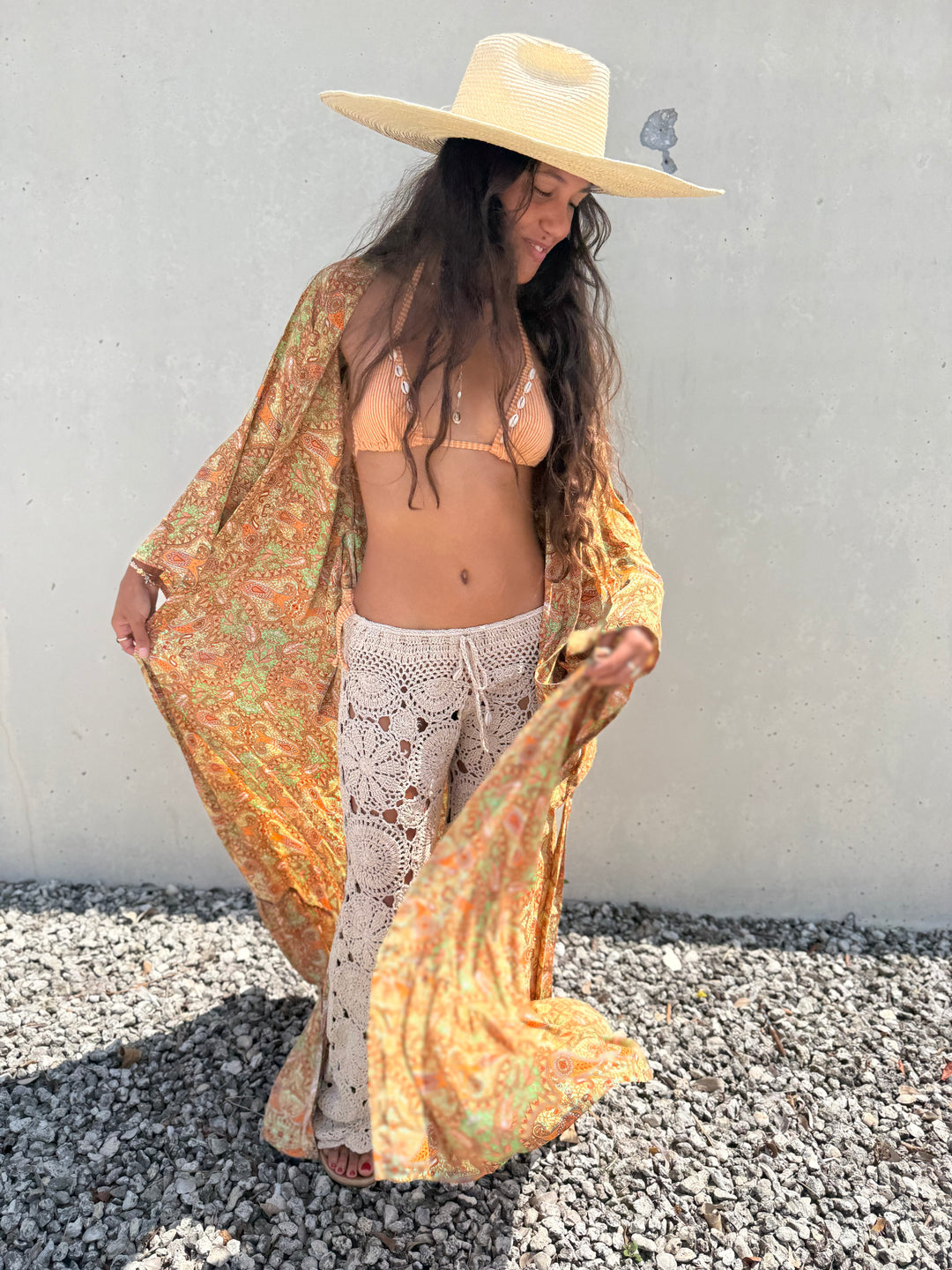 A woman wearing a bohemian-style outfit stands outdoors against a light gray wall on gravel. She is wearing a wide-brimmed straw hat, a sheer orange and green paisley kaftan, cream crochet boho pants, and a matching cream bikini top.