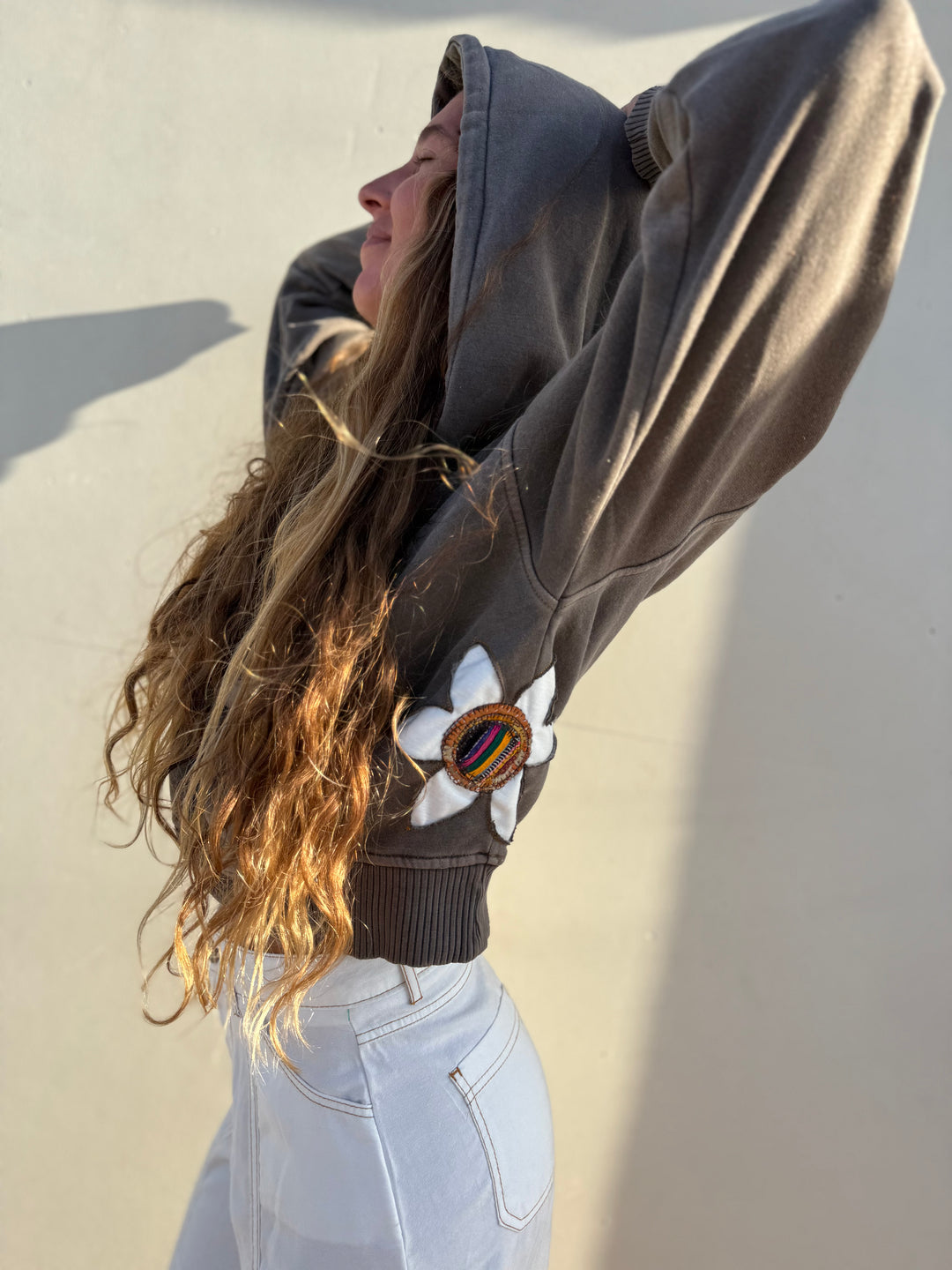 A woman wearing a dark brown oversized hoodie with a white flower patch and a colorful striped circular accent on the sleeve, paired with light wash jeans, posing against a neutral wall.