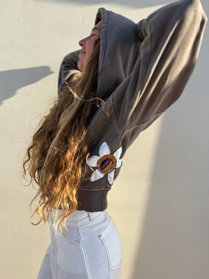 A woman wearing a dark brown oversized hoodie with a white flower patch and a colorful striped circular accent on the sleeve, paired with light wash jeans, posing against a neutral wall.