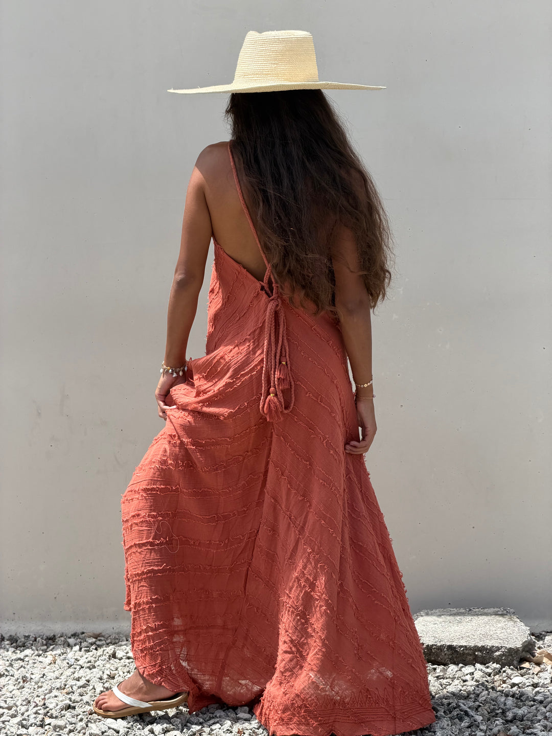 A woman viewed from behind wearing a long terracotta-colored linen maxi dress with a tie-back neckline, a wide-brimmed straw hat, and sandals, standing against a plain light gray wall on a gravel surface.
