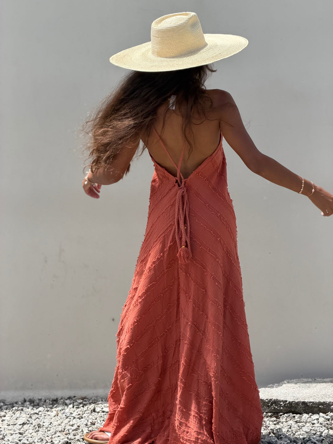 A woman wearing a wide-brimmed straw hat and a textured terracotta maxi dress with a tie-back back, photographed from behind against a plain white wall.
