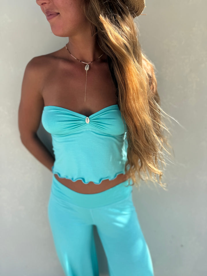A woman wearing a matching turquoise blue two-piece set consisting of a strapless ruched crop top with a scalloped hem and high-waisted leggings, shown against a plain light gray wall.