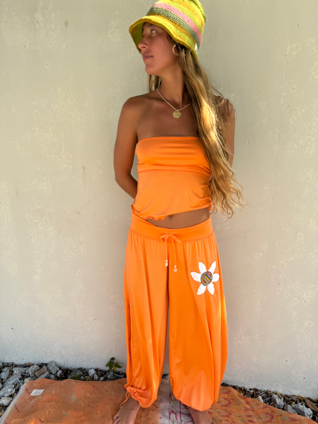 A woman wearing a bright orange two-piece lounge set consisting of a strapless crop top and wide-leg drawstring pants with a white flower graphic, paired with a yellow and pink striped sun hat, standing against a textured beige wall.