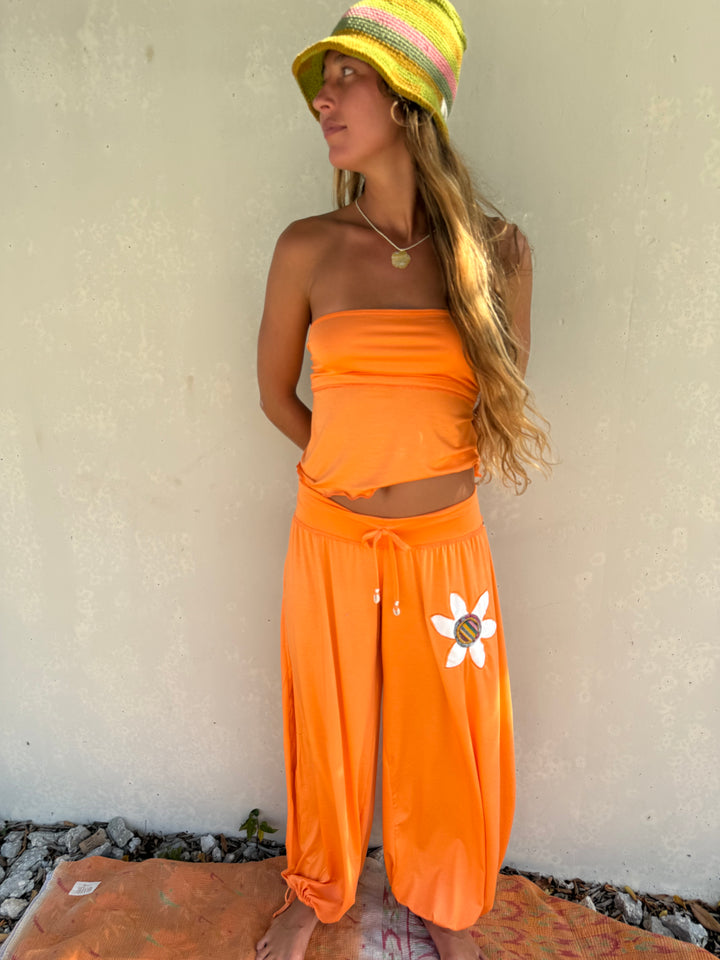A woman wearing a bright orange two-piece lounge set consisting of a strapless crop top and wide-leg drawstring pants with a white flower graphic, paired with a yellow and pink striped sun hat, standing against a textured beige wall.