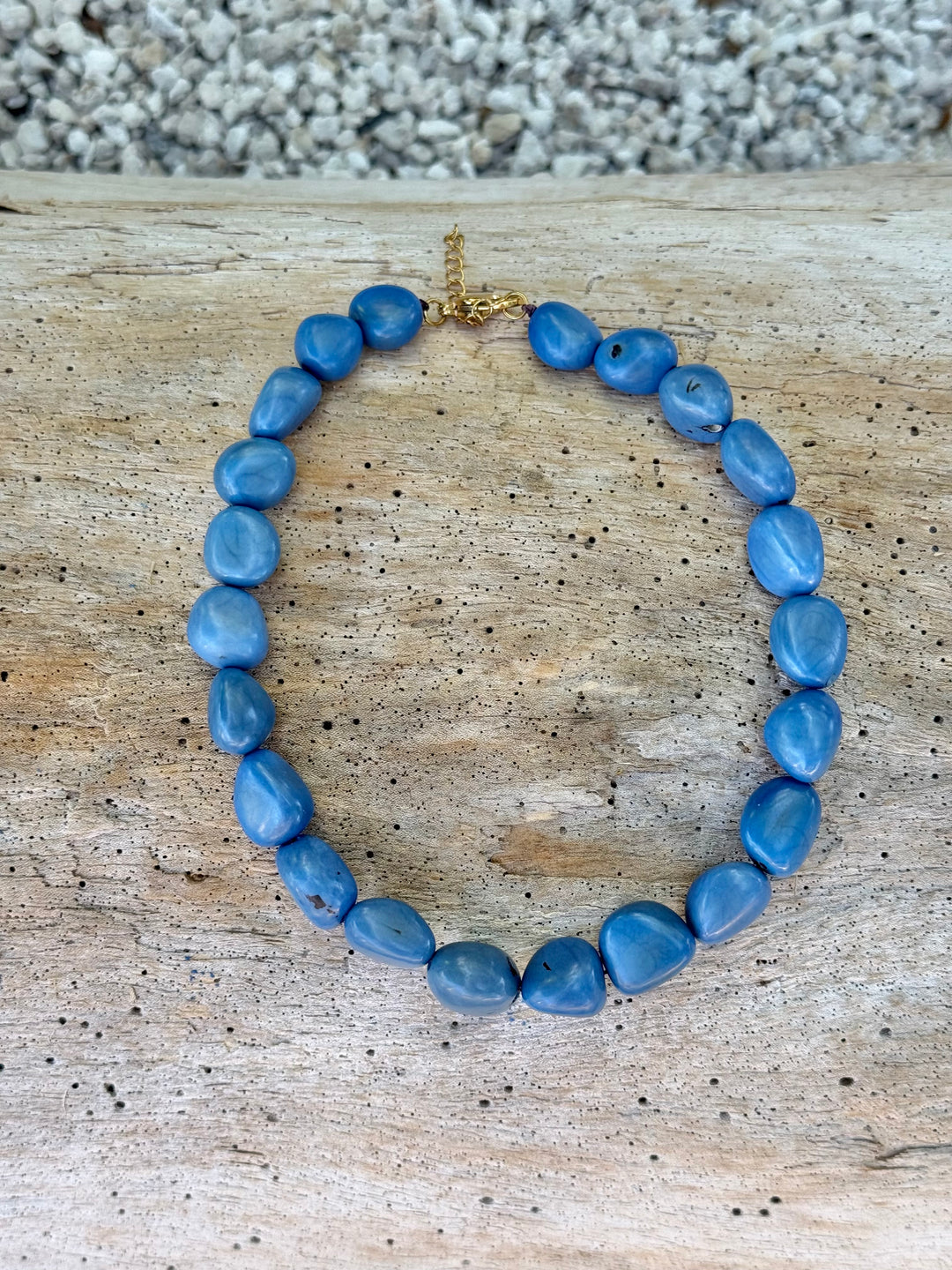 A beaded necklace made of irregular medium-blue stones arranged in an oval, with a small gold clasp at the top, photographed on a weathered wood surface with gravel in the background.