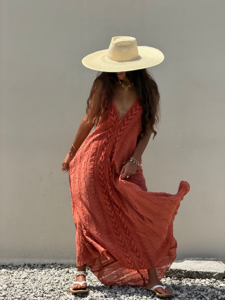 A woman wearing a wide-brimmed straw hat and a flowing terracotta-colored maxi dress with a cable-knit pattern, standing against a plain light gray wall on a gravel surface.