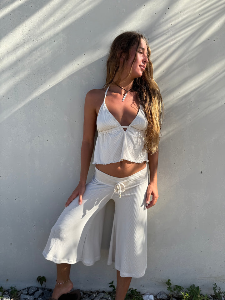 A woman posing outdoors against a textured white wall with dappled sunlight and shadows, wearing a white two-piece lounge set consisting of a halter crop top and wide-leg drawstring pants.