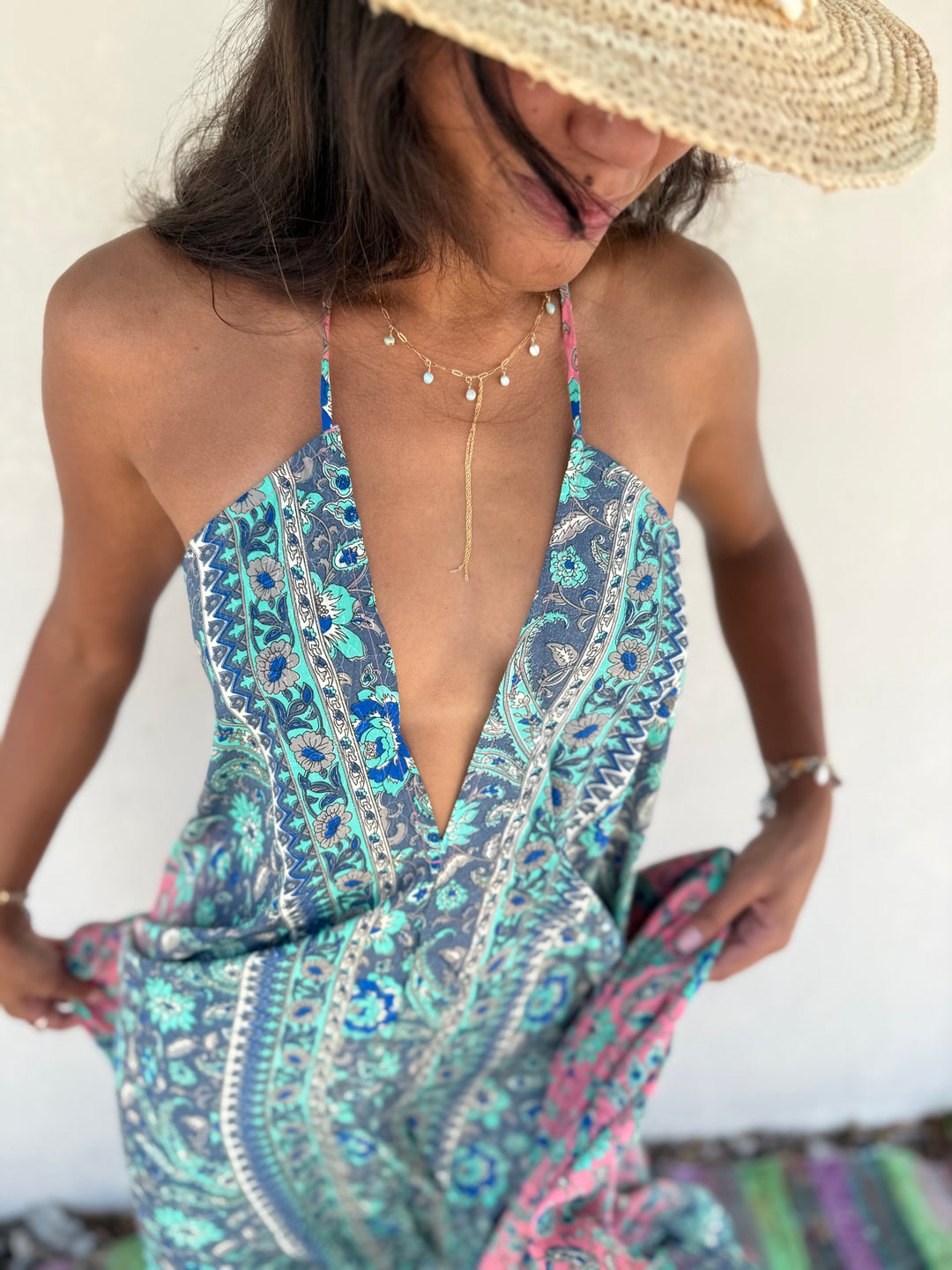 A woman wearing a bohemian-style halter maxi dress with a blue, teal, and pink paisley print, accessorized with a straw hat and gold necklaces.