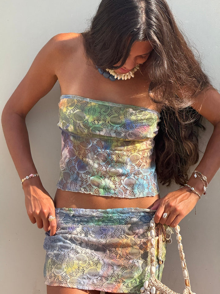 A woman wearing a matching two-piece set with a strapless tube top and wrap skirt in a multicolor floral lace pattern, accessorized with a chunky shell necklace and bracelets, posing against a neutral wall.