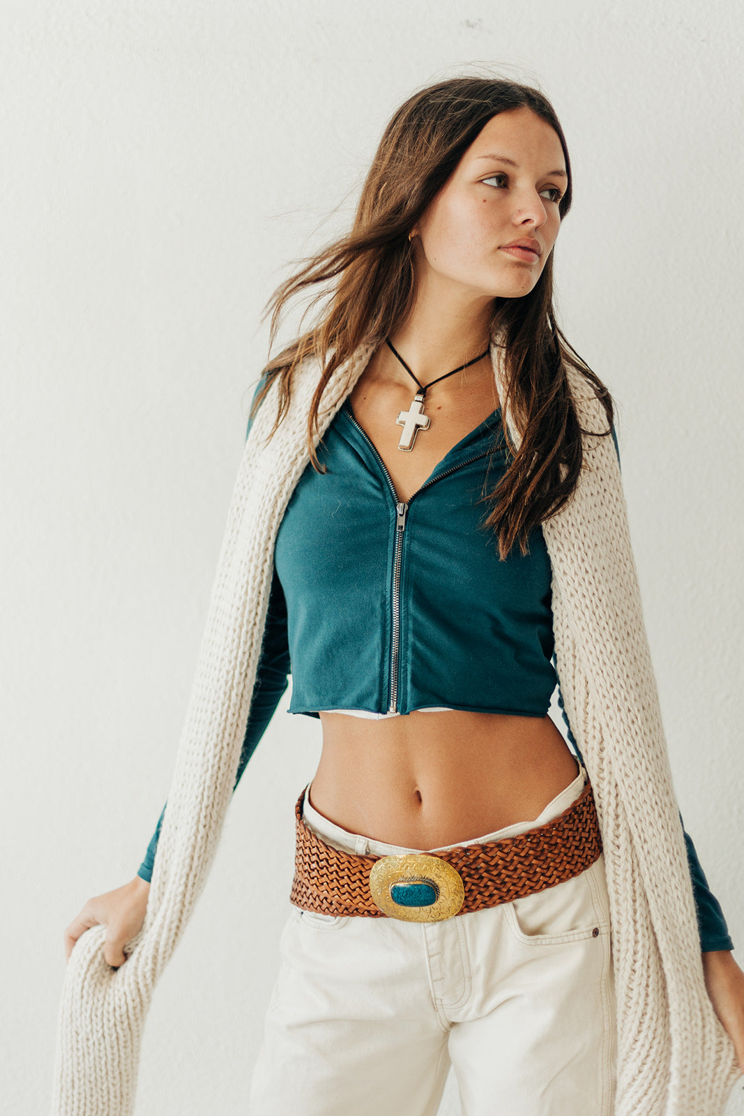 Woman wearing a green top, beige cardigan, and white pants with a decorative belt against a plain background