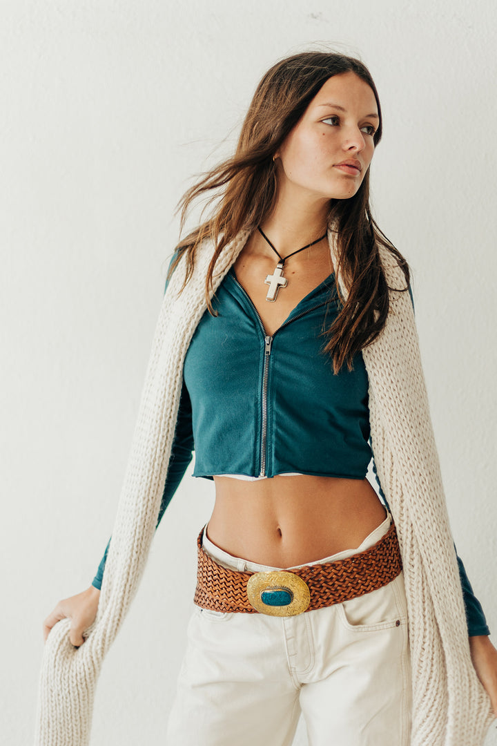 Woman wearing a green top, beige cardigan, and white pants with a decorative belt against a plain background