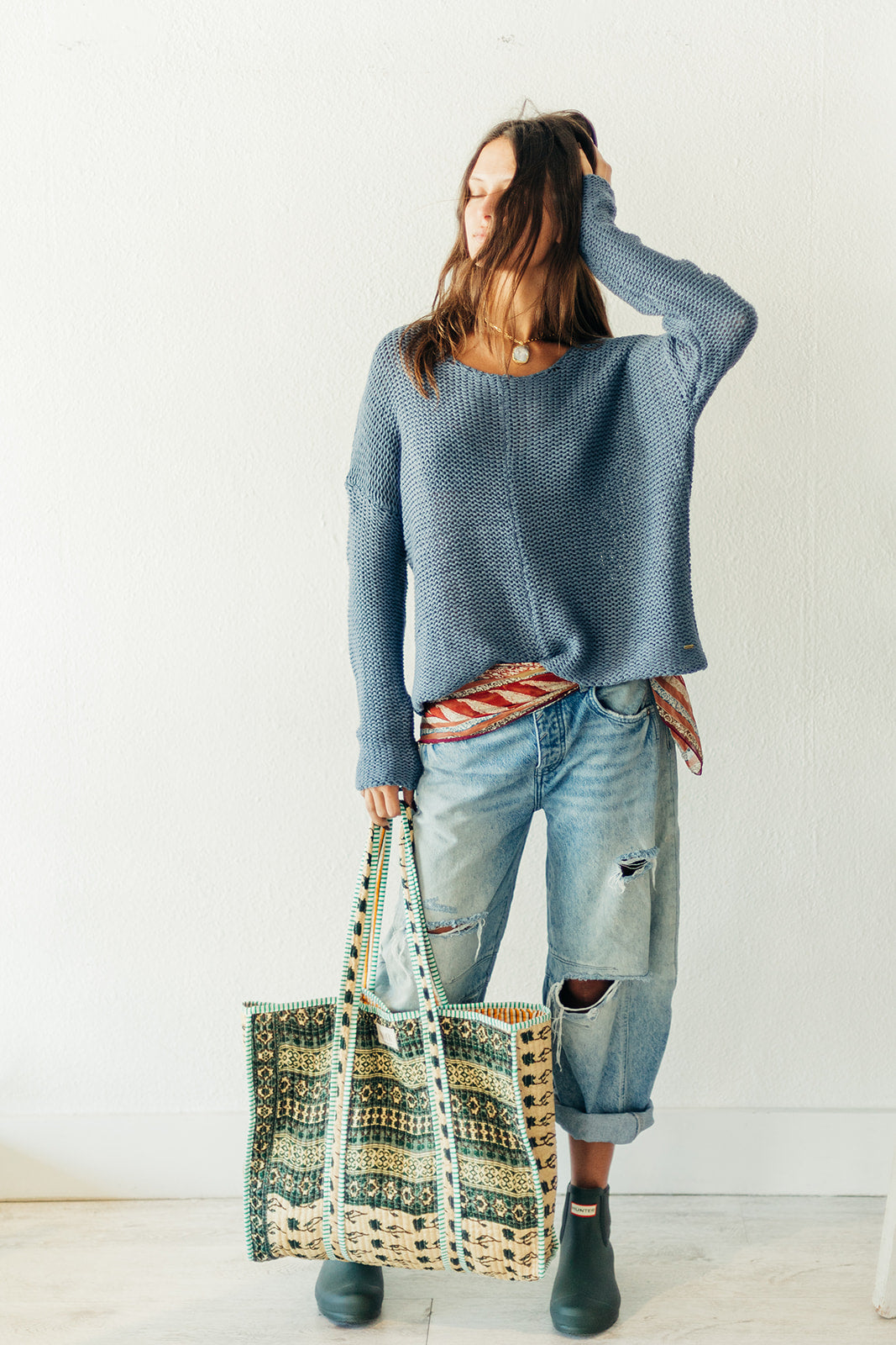 Woman wearing a blue sweater and jeans, holding a patterned bag against a white background