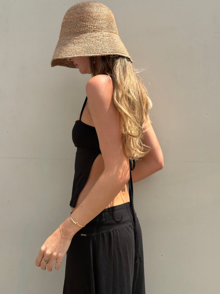 A woman wearing a natural straw bucket hat, a black cutout camisole, and black wide-leg pants, standing against a plain light gray wall.