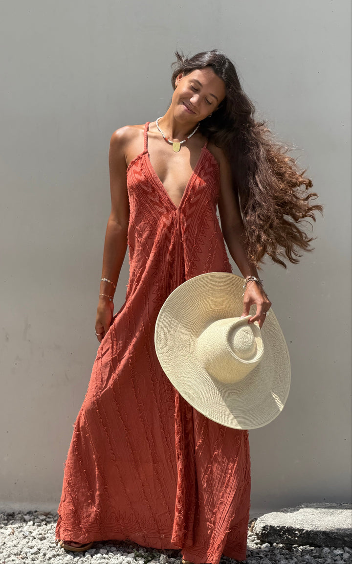A woman wearing a rust-orange maxi dress and holding a large beige straw hat, standing against a plain light gray wall on a gravel surface.
