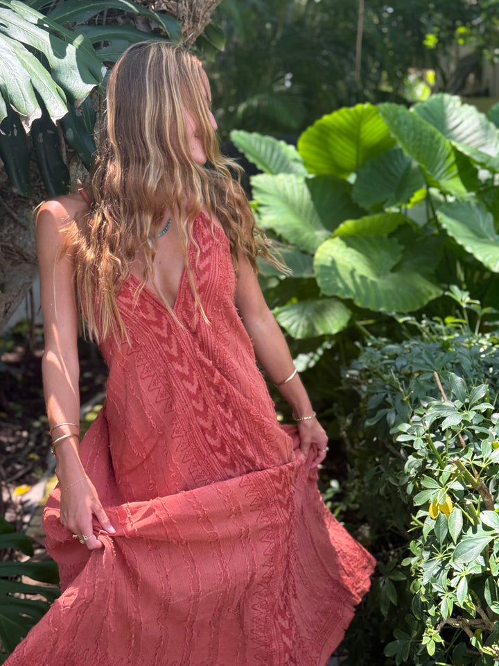 A woman wearing a textured coral pink sleeveless maxi dress, standing outdoors in a lush garden and holding out the skirt to show its flow.