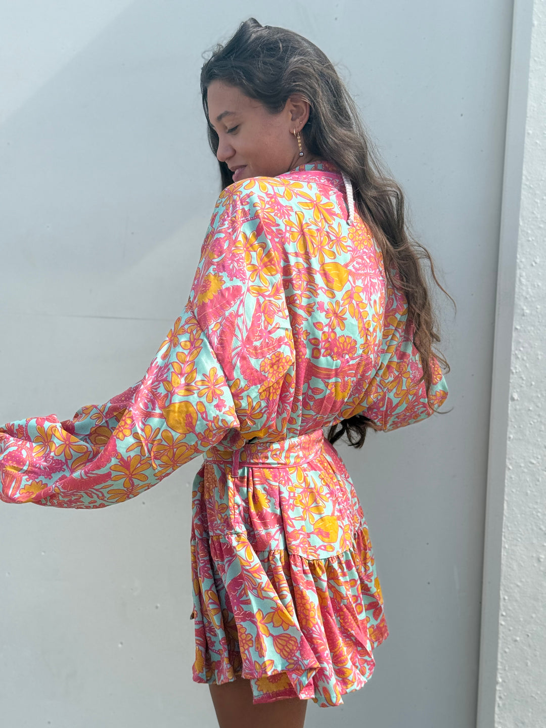 A woman wearing a colorful tropical print kimono-style dress, photographed from the back against a plain light gray wall.