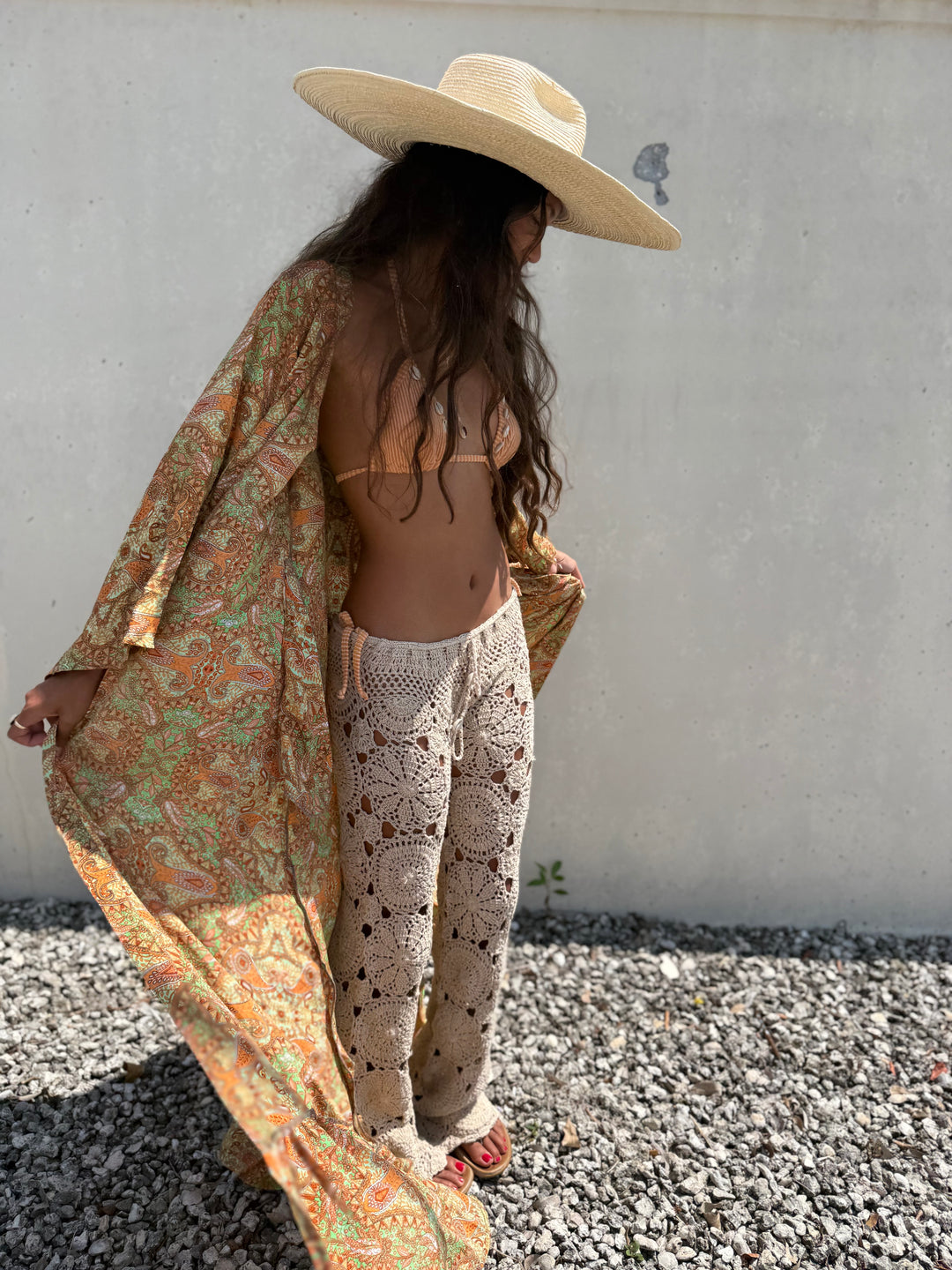 A woman stands outdoors against a light concrete wall on gravel, wearing a wide-brim straw hat, a loose patterned orange and green cover-up, crochet boho pants, and red sandals.