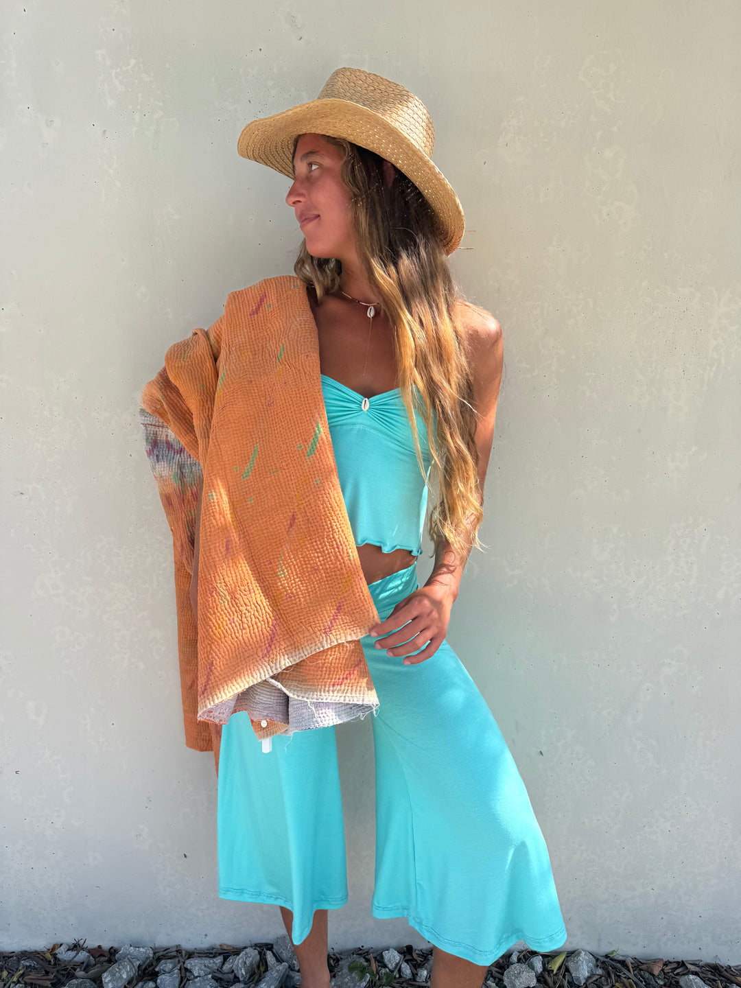 A woman wearing a straw hat, a turquoise two-piece set, and holding an orange patterned wrap against a textured off-white wall.