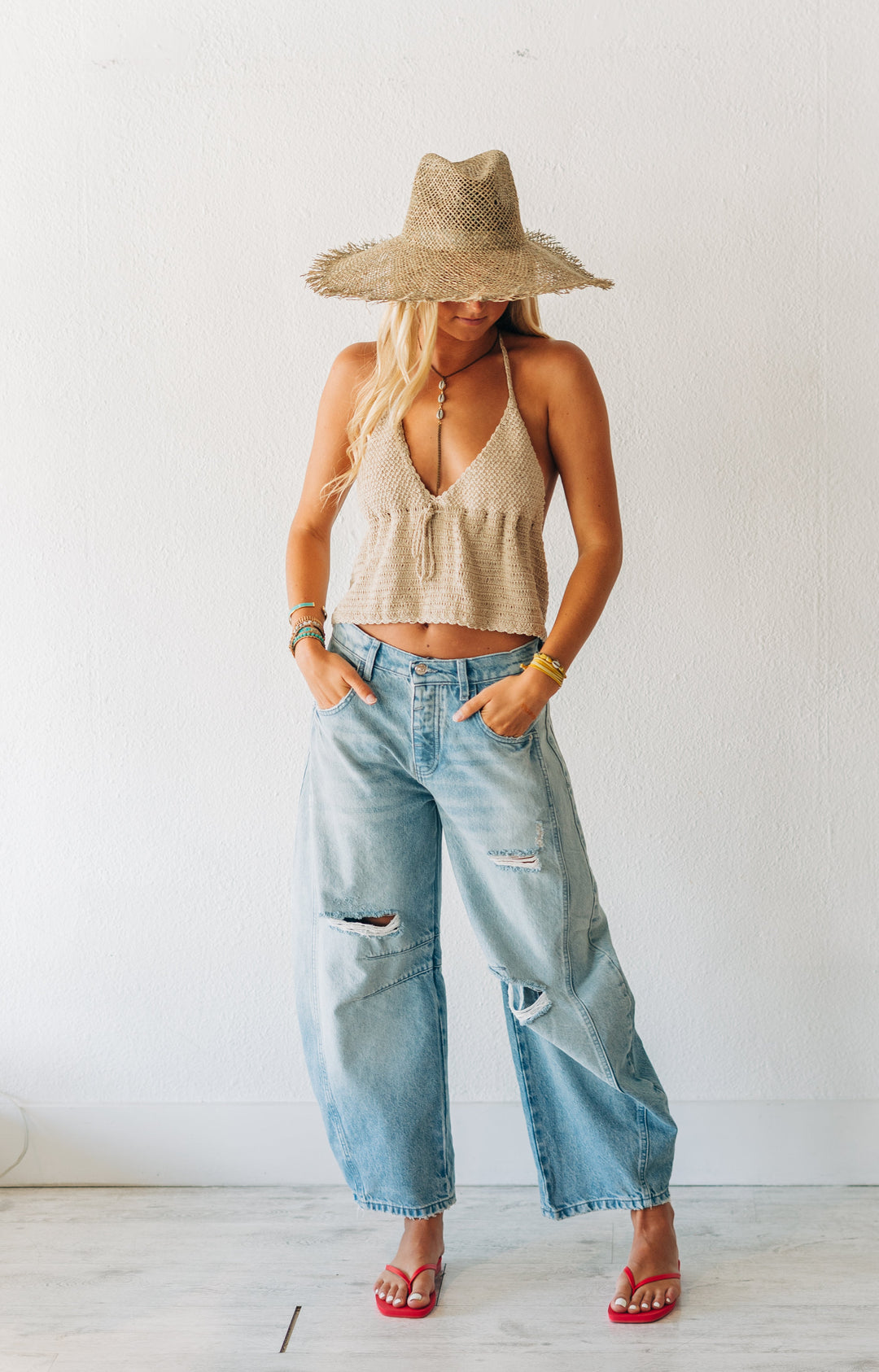 Woman wearing a beige top, wide-brimmed hat, and blue jeans in a minimal indoor setting.