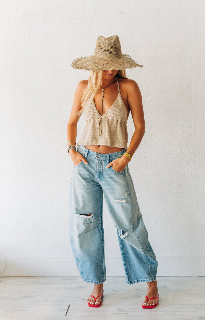 Woman wearing a beige top, wide-brimmed hat, and blue jeans in a minimal indoor setting.
