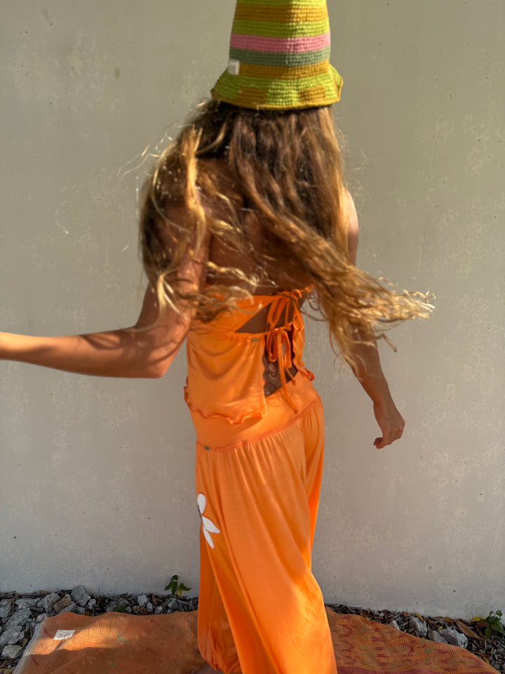 A woman wearing a bright orange strapless maxi dress and a green striped bucket hat, photographed from behind against a light gray stucco wall.
