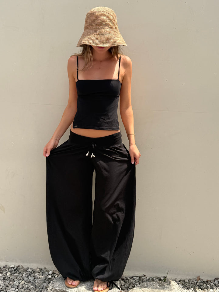 A woman wearing a beige straw bucket hat, a black spaghetti-strap crop top, and black wide-leg pants stands against a plain light gray wall.