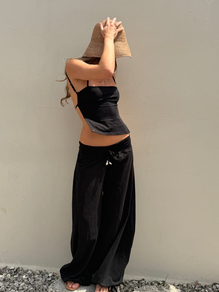 A woman wearing a black camisole top and matching black wide-leg pants, standing against a plain beige wall outdoors while holding a straw hat over her face.